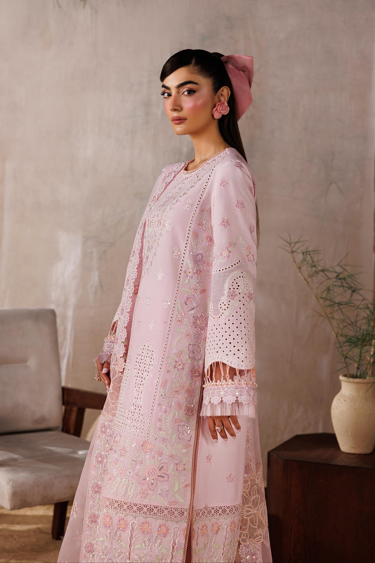 Woman in a pink traditional outfit standing against a textured wall with a plant and chair in the background.