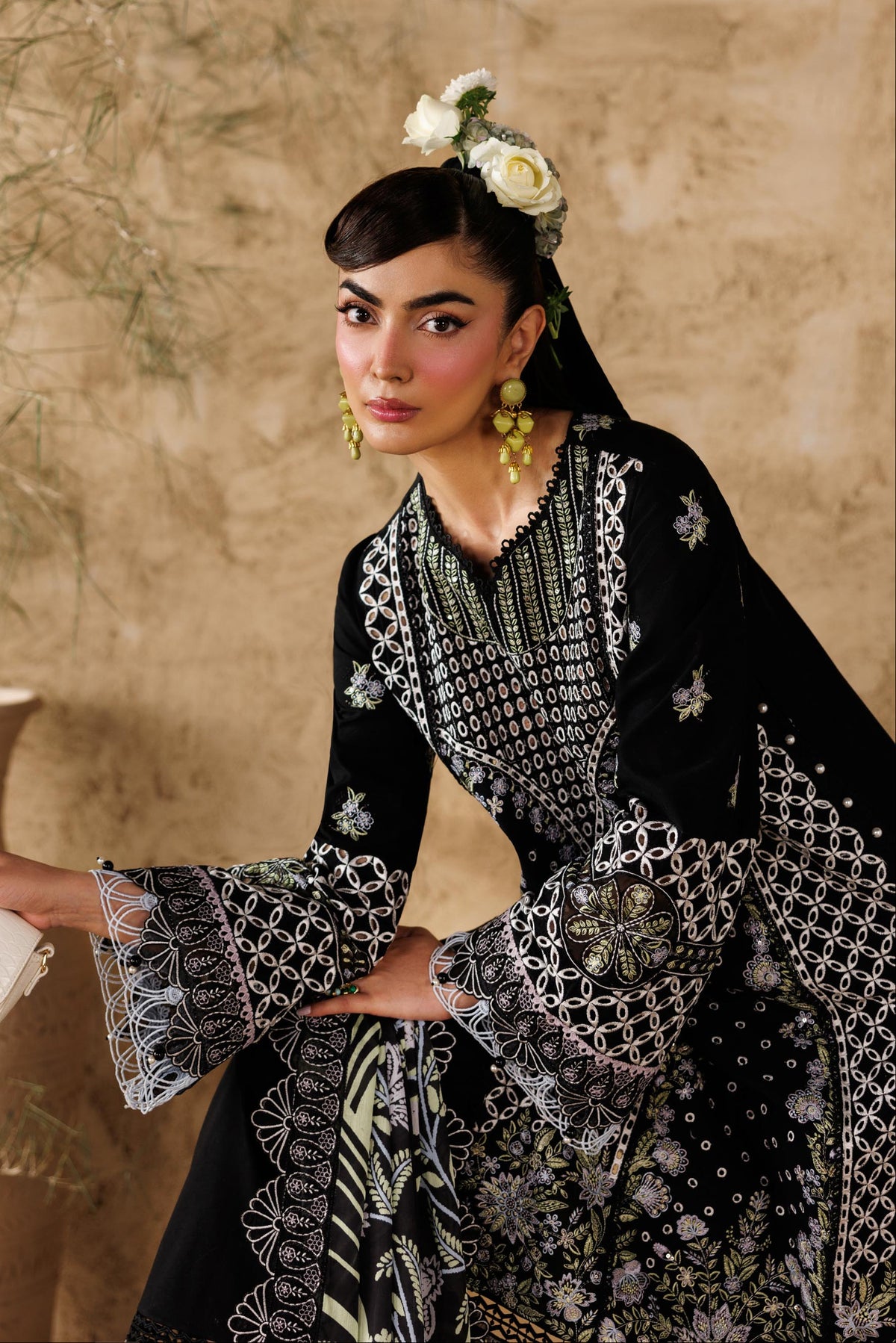 Woman in a black and white patterned dress with a floral headpiece, holding a white clutch, against a beige wall.