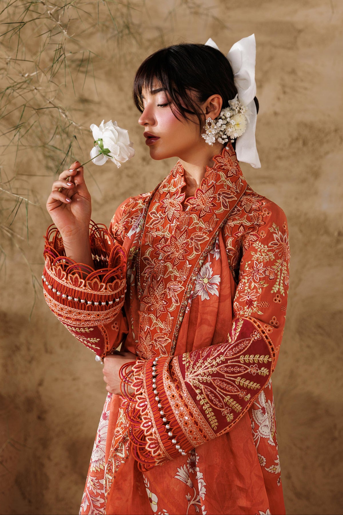 Woman in a traditional red and gold outfit holding a white flower against a beige wall.