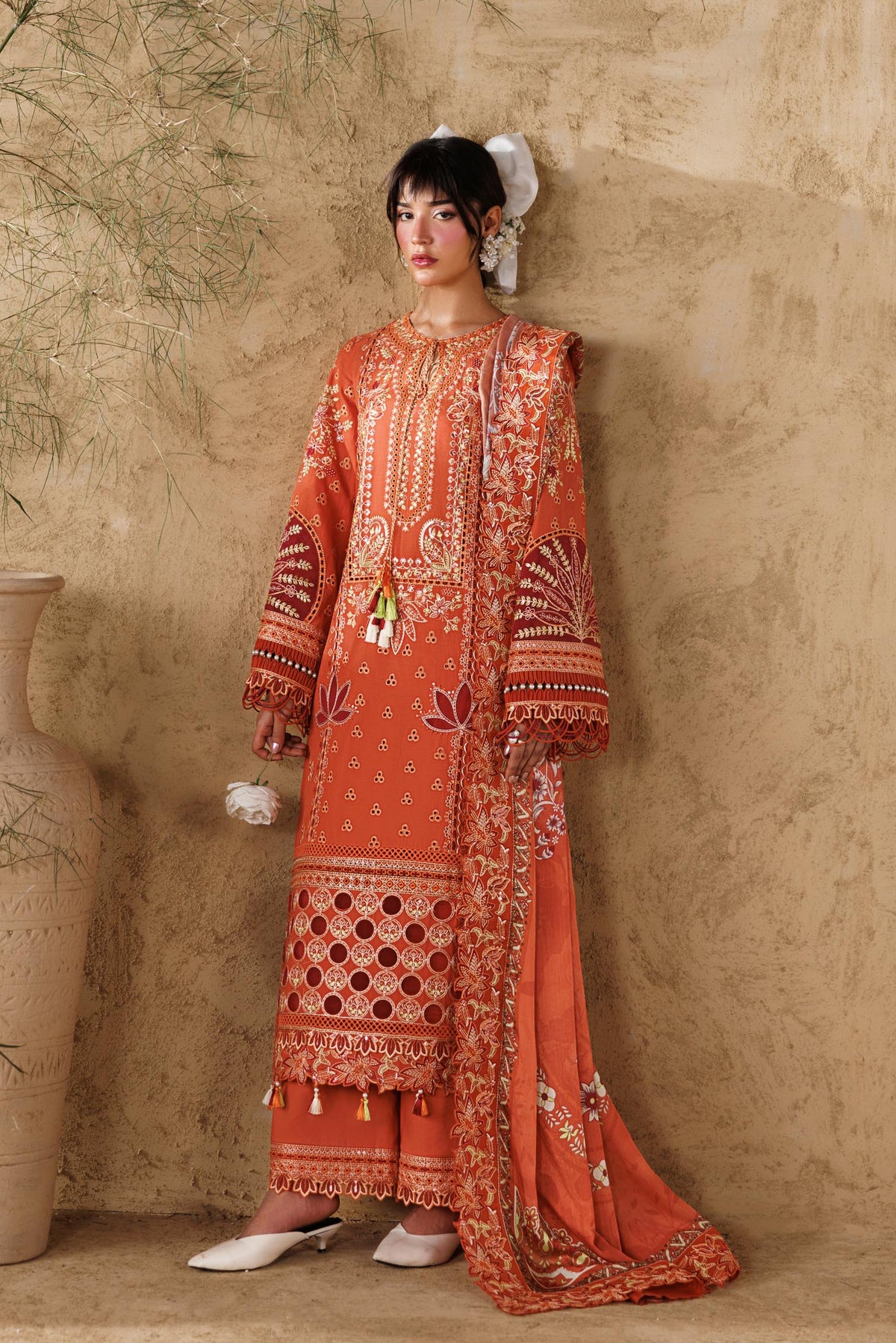 Woman in an orange embroidered outfit standing against a textured beige wall with decorative vases and plants.