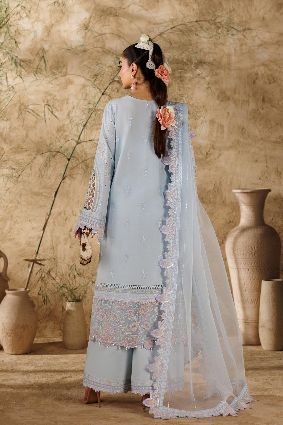 Woman in a light blue traditional outfit standing in a rustic setting with vases and plants.