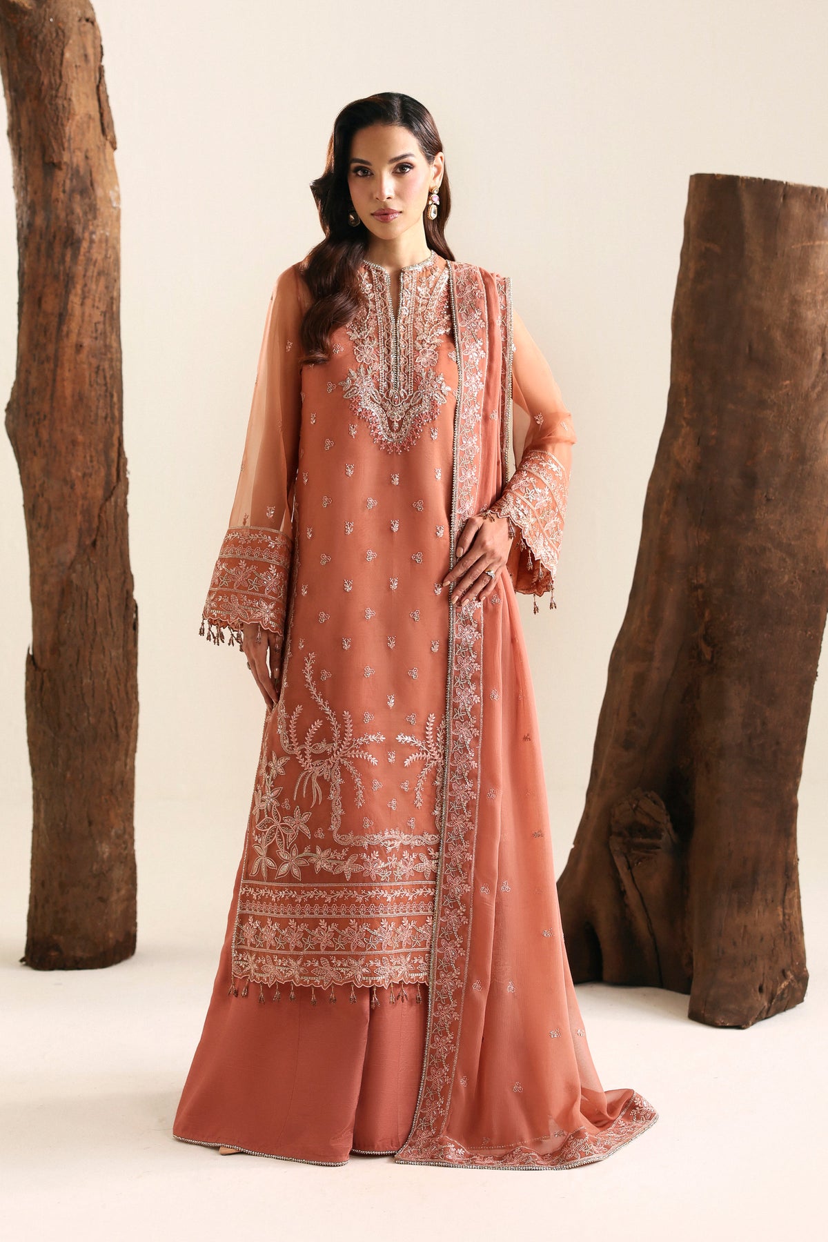 A woman models a peach-colored traditional South Asian outfit with intricate silver embroidery, consisting of a long kurta, matching pants, and a dupatta, set against a studio backdrop with rustic wooden logs.