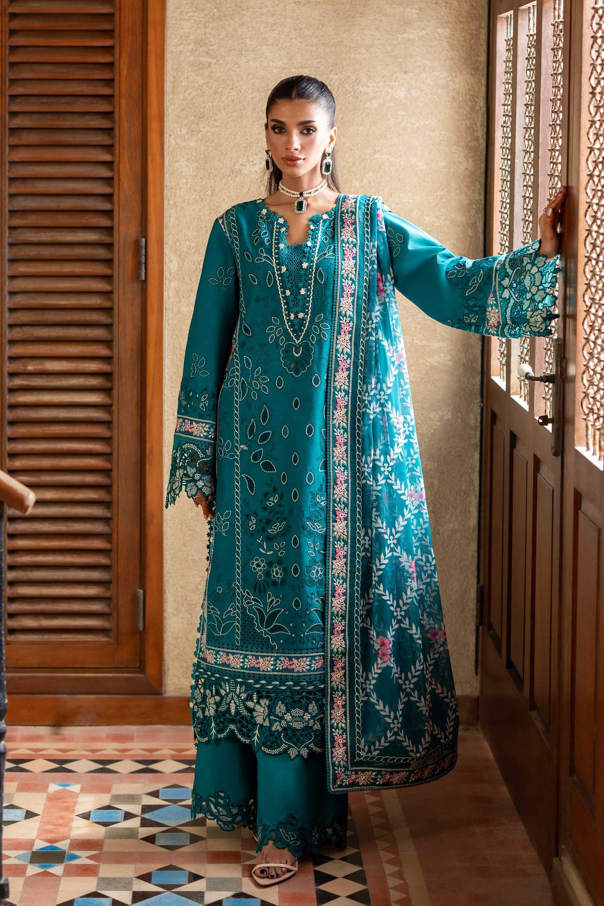 Woman in a teal embroidered traditional outfit standing in a room with wooden doors and a patterned floor.
