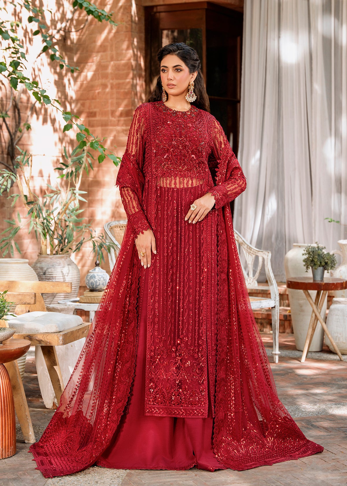 A woman wearing a red, netted, embroidered dress with long sleeves and a maxi skirt, standing in a room with curtains behind.