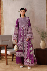 Woman in a purple embroidered outfit standing in front of a white curtain with a wooden frame.