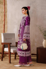 Woman in a purple traditional outfit standing in a room with a white curtain and wooden furniture.