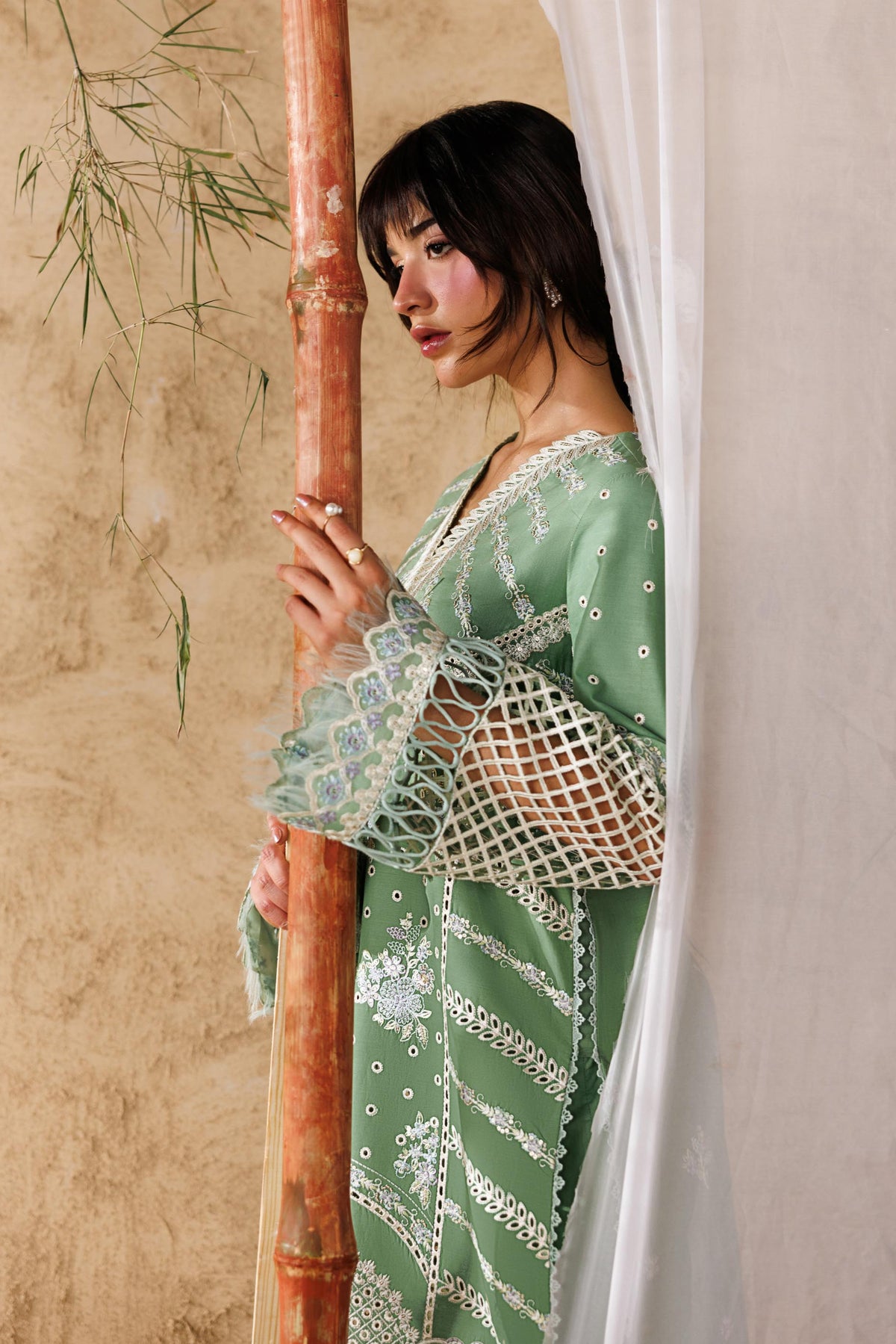 Woman in a green dress standing behind a bamboo stick against a beige wall.