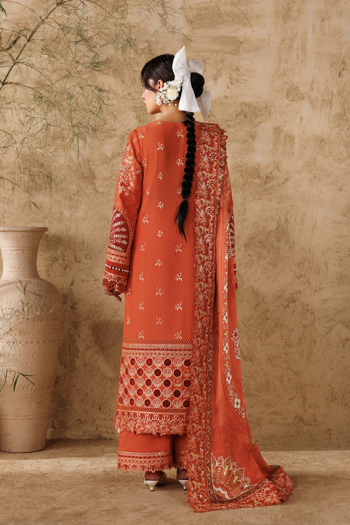 Woman in a traditional red embroidered outfit standing against a textured beige wall with decorative vases.