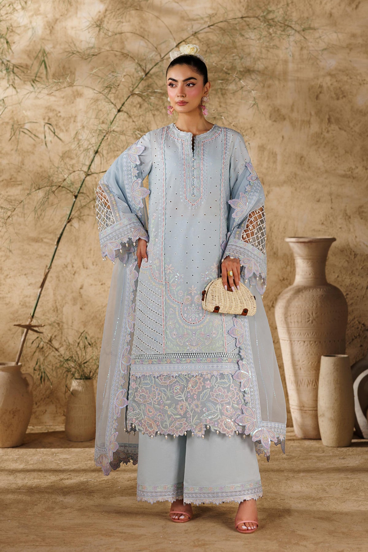 Woman in a light blue embroidered outfit standing in a rustic setting with vases and plants.
