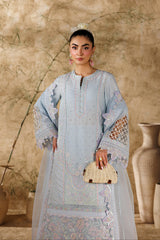 Woman in a light blue embroidered dress holding a beige handbag against a textured wall.