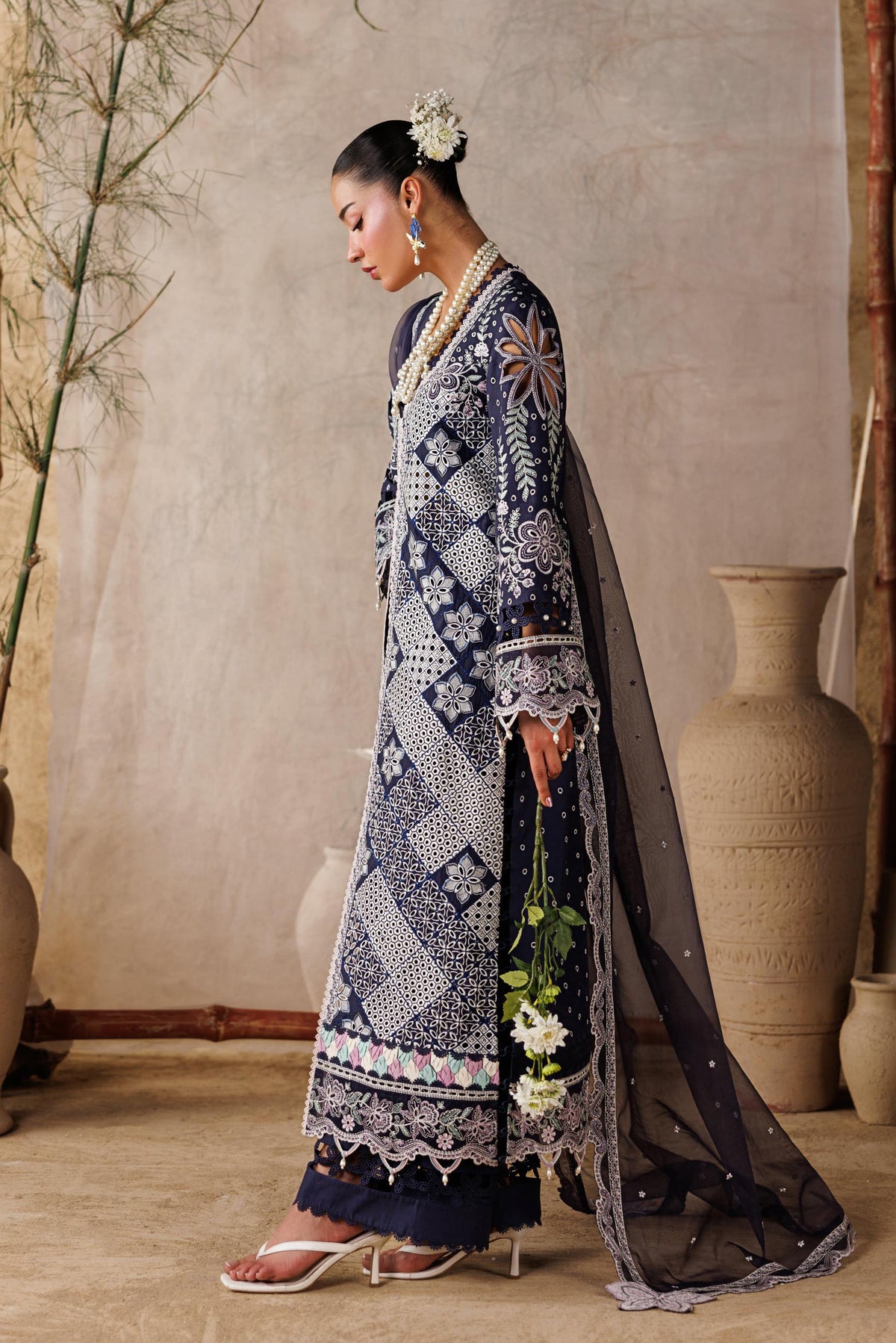 Woman in a traditional outfit standing in a room with decorative vases and plants.
