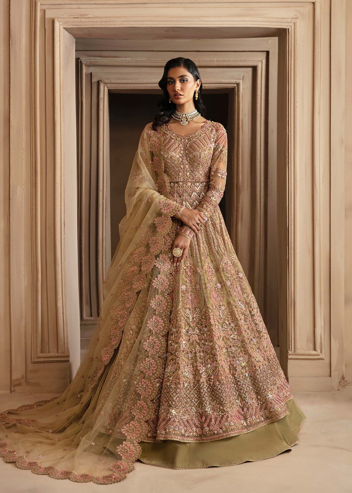 A woman wearing a beige and gold embroidered dress with netting and lehenga, standing in a doorway.