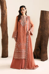 A woman models a peach-colored traditional South Asian outfit with intricate silver embroidery, consisting of a long kurta, matching pants, and a dupatta, set against a studio backdrop with rustic wooden logs.