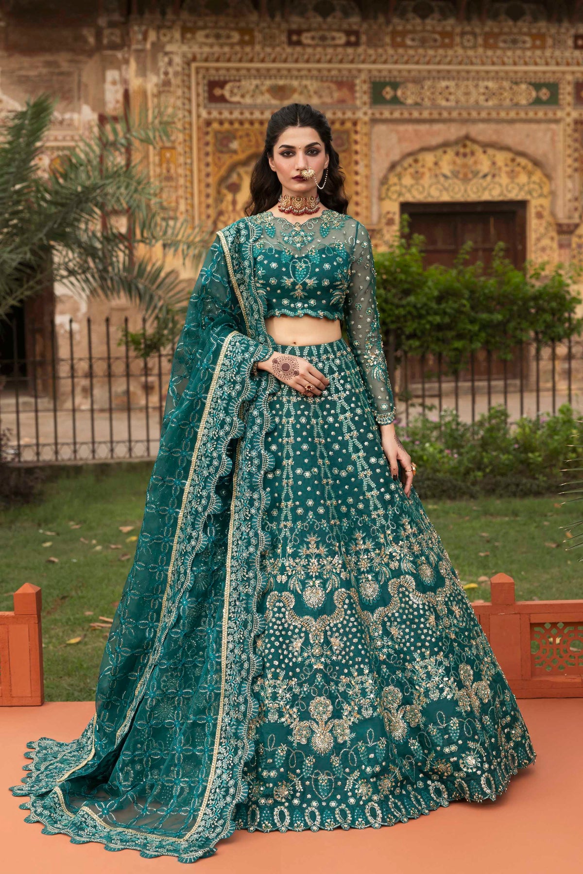 Woman in a traditional teal embroidered outfit standing outdoors with decorative background