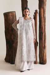 Woman in a white traditional outfit standing next to large wooden logs on a plain background