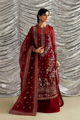 A woman modeling a deep maroon red traditional Indian outfit with intricate gold embroidery, featuring a long kurta, matching pants, and a sheer dupatta, posed against a silver draped backdrop.
