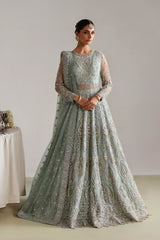 A woman modeling a heavily embroidered light sage green anarkali gown with intricate gold and silver beadwork, set against a neutral studio background.