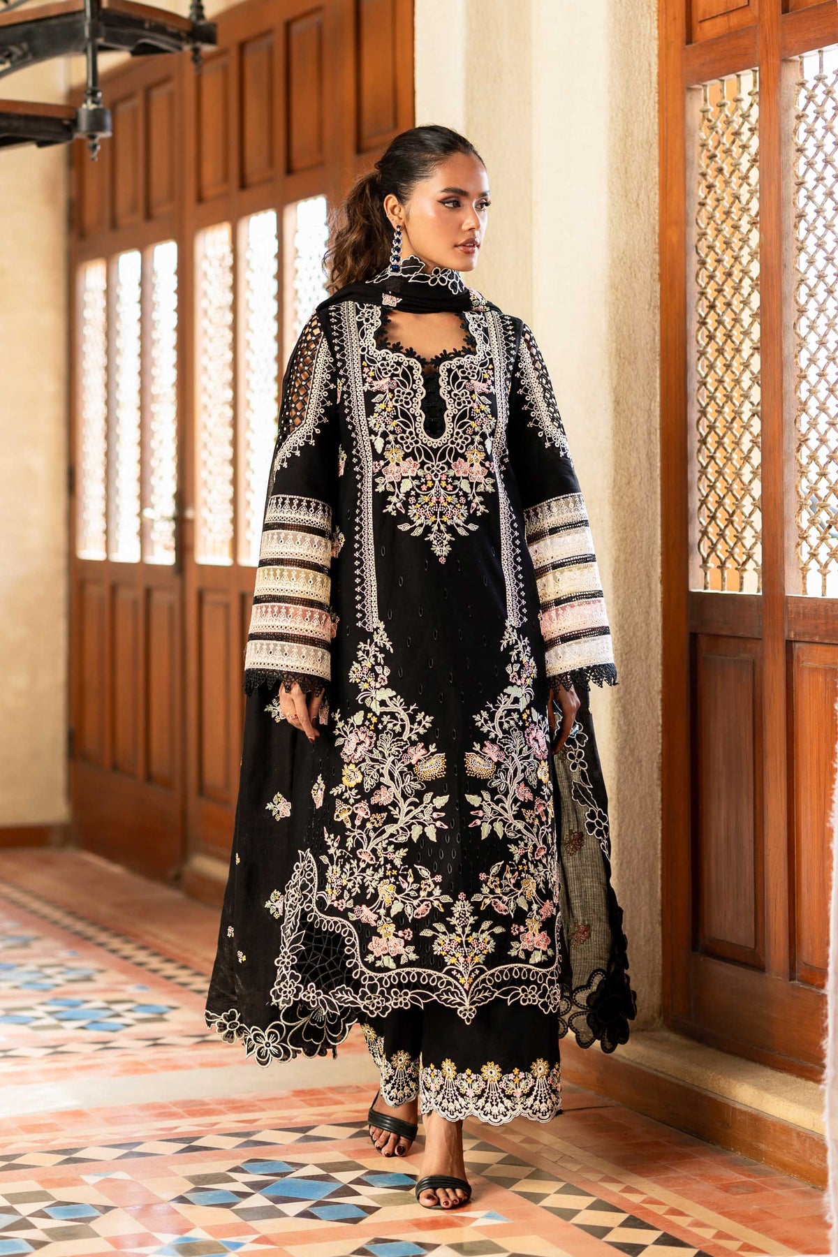 Woman in a black and white embroidered traditional outfit standing in a room with wooden doors and patterned floor.