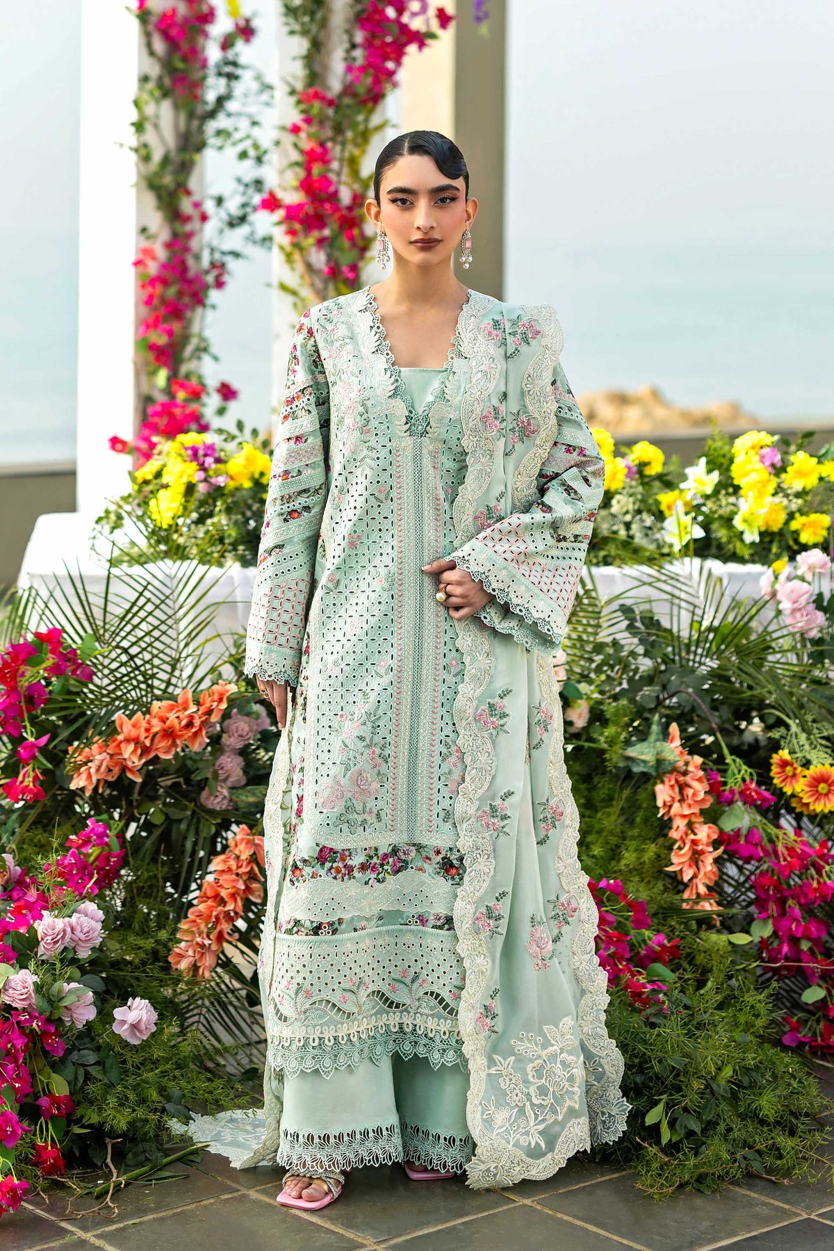 Woman in a light blue embroidered outfit standing in front of floral decorations.