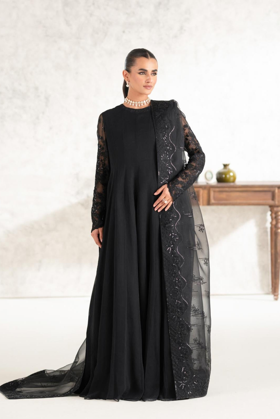 A woman wearing a black maxi dress with long sleeves and floral patterns, standing beside a wooden table.
